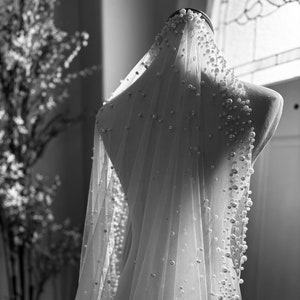May include: A white bridal veil with pearl embellishments. The veil is draped over a mannequin and has a soft, flowing texture.