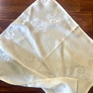 May include: A white linen napkin with a subtle floral pattern. The napkin is folded in a triangular shape and is lying on a wooden surface.