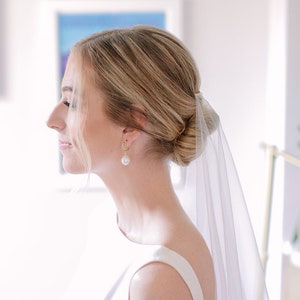 Pode incluir: Uma mulher em um vestido branco sem mangas com um coque baixo e um véu transparente. Ela está usando brincos de pérolas. O fundo é uma sala iluminada com obras de arte emolduradas.