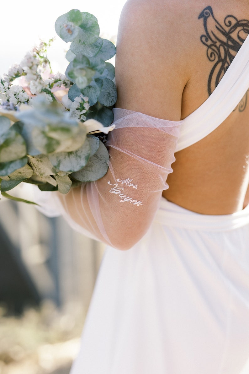 May include: A bride's arm is adorned with a sheer, light pink sleeve, embroidered with "Mrs. Nguyen" in white script. She holds a bouquet of white flowers and eucalyptus leaves. The back of a white dress is visible.