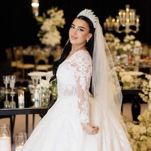 K&ouml;nnte beinhalten: Eine Braut in einem wei&szlig;en Hochzeitskleid mit langen Spitzen&auml;rmeln und einem weiten Rock. Sie tr&auml;gt ein Diadem und einen Schleier. Der Hintergrund zeigt Blumenschmuck und Kerzen, was eine festliche Atmosph&auml;re schafft.