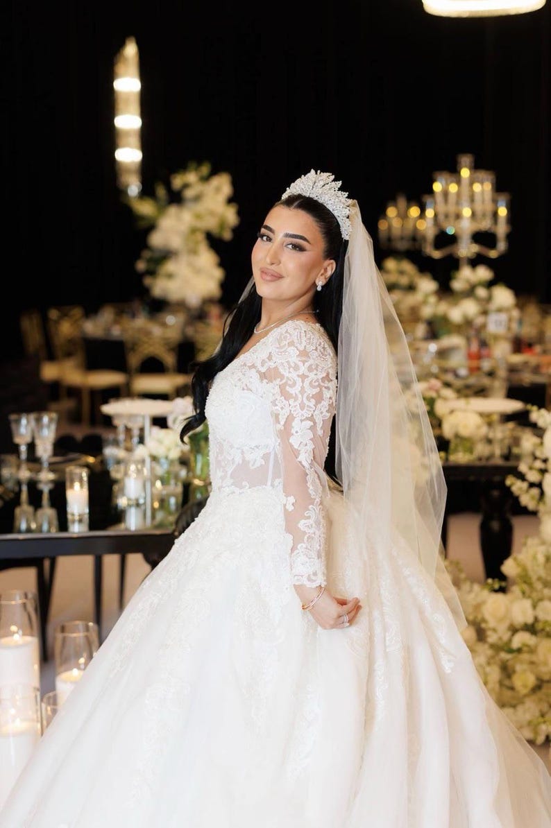 K&ouml;nnte beinhalten: Eine Braut in einem wei&szlig;en Hochzeitskleid mit langen Spitzen&auml;rmeln und einem weiten Rock. Sie tr&auml;gt ein Diadem und einen Schleier. Der Hintergrund zeigt Blumenschmuck und Kerzen, was eine festliche Atmosph&auml;re schafft.