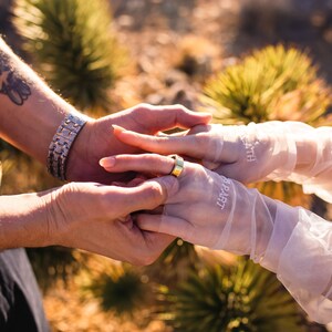 May include: A close-up shot of two sets of hands, one wearing a gold ring. One hand is wearing a silver bracelet. The other hand is wearing sheer white gloves with the words "PART" and "DEATH" printed on them. The background is blurred.
