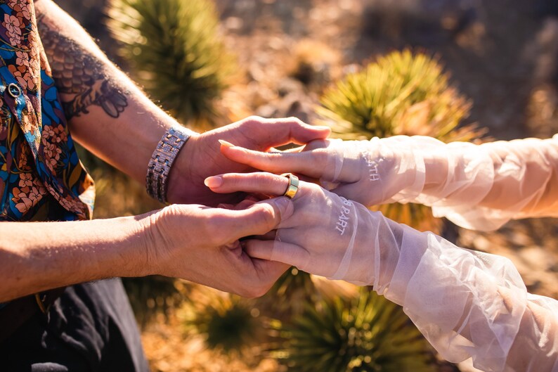 May include: A close-up shot of two sets of hands, one wearing a gold ring. One hand is wearing a silver bracelet. The other hand is wearing sheer white gloves with the words "PART" and "DEATH" printed on them. The background is blurred.