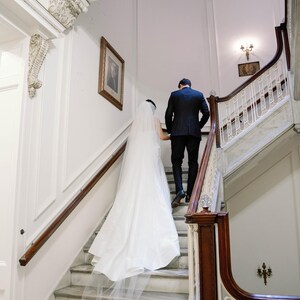 K&ouml;nnte beinhalten: Ein Paar steigt eine Marmortreppe in einem historischen Geb&auml;ude hinauf. Die Braut tr&auml;gt ein wei&szlig;es Brautkleid mit langem Schleier, und der Br&auml;utigam ist in einem schwarzen Anzug. Die Treppe hat ein Holzgel&auml;nder und verzierte Details.