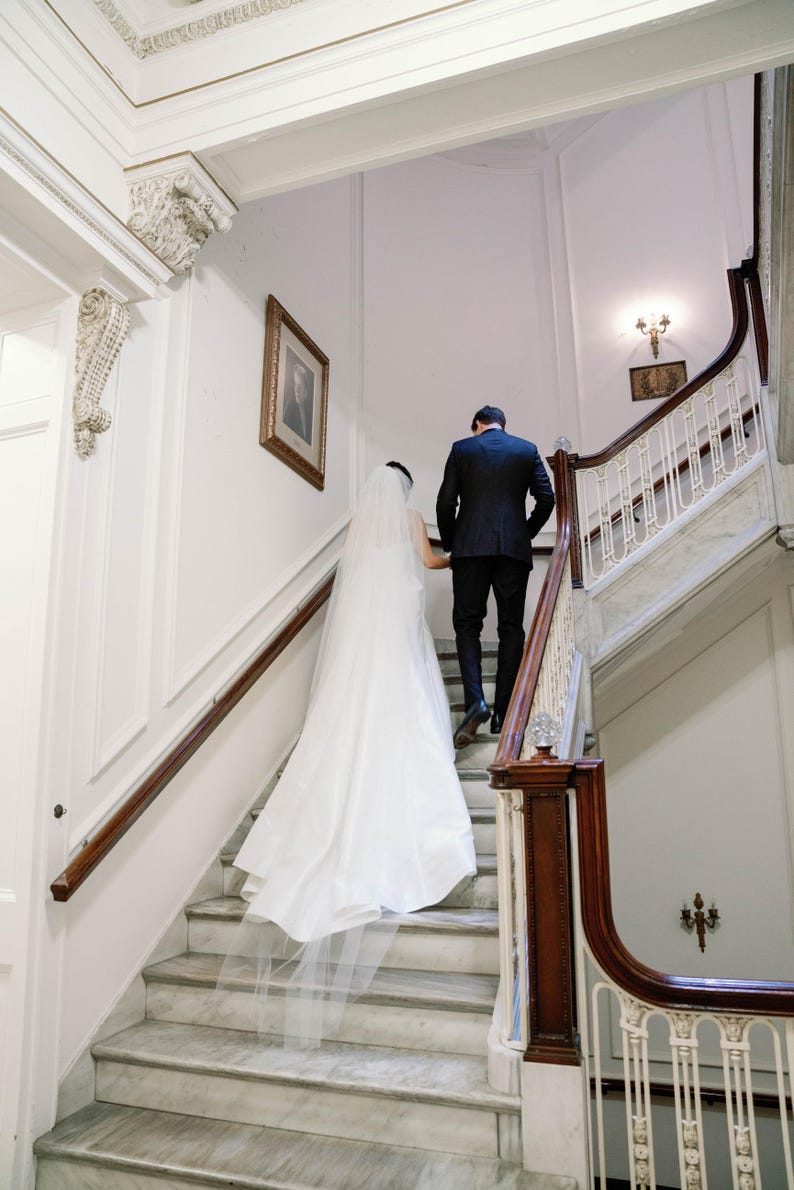 K&ouml;nnte beinhalten: Ein Paar steigt eine Marmortreppe in einem historischen Geb&auml;ude hinauf. Die Braut tr&auml;gt ein wei&szlig;es Brautkleid mit langem Schleier, und der Br&auml;utigam ist in einem schwarzen Anzug. Die Treppe hat ein Holzgel&auml;nder und verzierte Details.