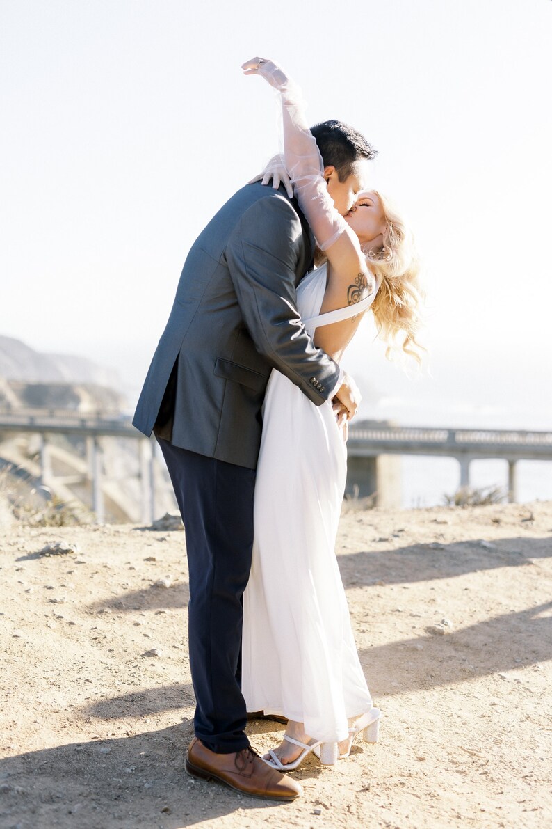 May include: A couple embraces and kisses outdoors. The person on the left wears a gray suit jacket, navy pants, and brown shoes. The person on the right wears a white dress and white heels. The person on the right has blonde hair and a tattoo on their arm.