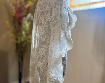 May include: An off-white lace veil featuring an intricate floral pattern throughout the fabric. The veil has a scalloped edge with delicate detailing and is draped over a mannequin, showcasing the semi-sheer texture and the detailed embroidery.