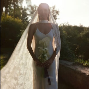 May include: A bride wearing a white satin wedding dress with a lace veil and a bouquet of white calla lilies.