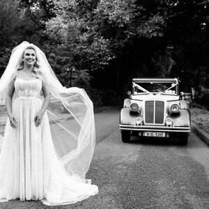 K&ouml;nnte beinhalten: Eine Braut in einem wei&szlig;en Hochzeitskleid und Schleier steht auf einer Stra&szlig;e, im Hintergrund ein Oldtimer. Das Bild ist in Schwarzwei&szlig;.