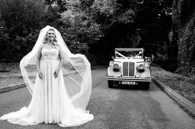 K&ouml;nnte beinhalten: Eine Braut in einem wei&szlig;en Hochzeitskleid und Schleier steht auf einer Stra&szlig;e, im Hintergrund ein Oldtimer. Das Bild ist in Schwarzwei&szlig;.
