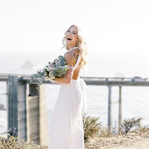 May include: A woman in a white jumpsuit laughs, holding a bouquet of flowers. The open-back jumpsuit has wide legs and a halter-style top. The background features a bridge and the ocean, with a sunny, outdoor setting.