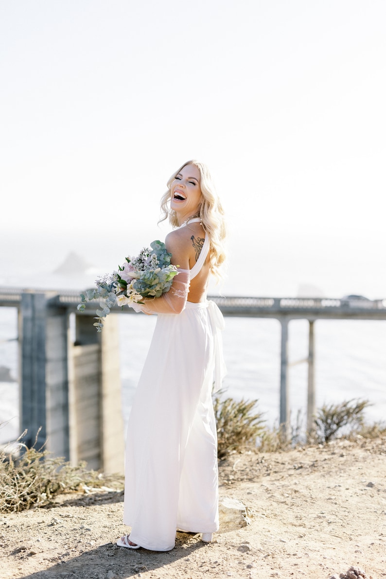 May include: A woman in a white jumpsuit laughs, holding a bouquet of flowers. The open-back jumpsuit has wide legs and a halter-style top. The background features a bridge and the ocean, with a sunny, outdoor setting.