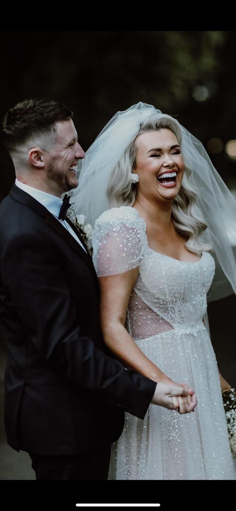 K&ouml;nnte beinhalten: Braut und Br&auml;utigam teilen einen freudigen Moment und lachen zusammen. Die Braut tr&auml;gt ein wei&szlig;es, verziertes Hochzeitskleid mit Schleier. Der Br&auml;utigam tr&auml;gt einen schwarzen Smoking. Das Bild zeigt eine Hochzeitsfeier.