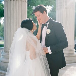 K&ouml;nnte beinhalten: Eine Braut in einem wei&szlig;en Hochzeitskleid und Schleier ber&uuml;hrt das Gesicht eines Br&auml;utigams in einem schwarzen Smoking. Der Br&auml;utigam tr&auml;gt eine schwarze Fliege und eine wei&szlig;e Blume am Revers. Sie stehen in der N&auml;he gro&szlig;er wei&szlig;er S&auml;ulen.