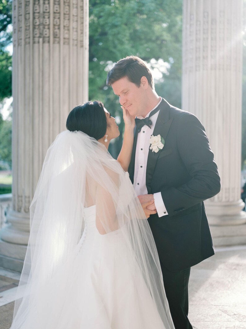 K&ouml;nnte beinhalten: Eine Braut in einem wei&szlig;en Hochzeitskleid und Schleier ber&uuml;hrt das Gesicht eines Br&auml;utigams in einem schwarzen Smoking. Der Br&auml;utigam tr&auml;gt eine schwarze Fliege und eine wei&szlig;e Blume am Revers. Sie stehen in der N&auml;he gro&szlig;er wei&szlig;er S&auml;ulen.