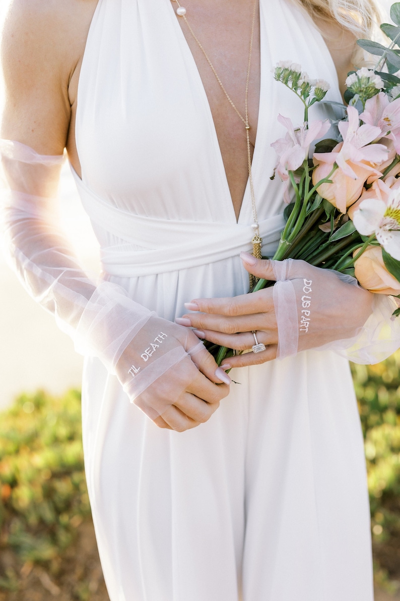 May include: A white wedding dress with a plunging neckline. The bride is wearing sheer gloves with the words "TIL DEATH" and "DO US PART" embroidered on them. She holds a bouquet of flowers and wears a gold necklace.