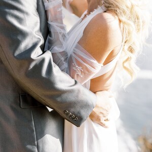 May include: A wedding scene featuring a gray suit jacket with four buttons and a white dress. The bride wears sheer gloves with the text "Mrs. Longton" embroidered on them. A diamond ring is visible. The background is blurred.