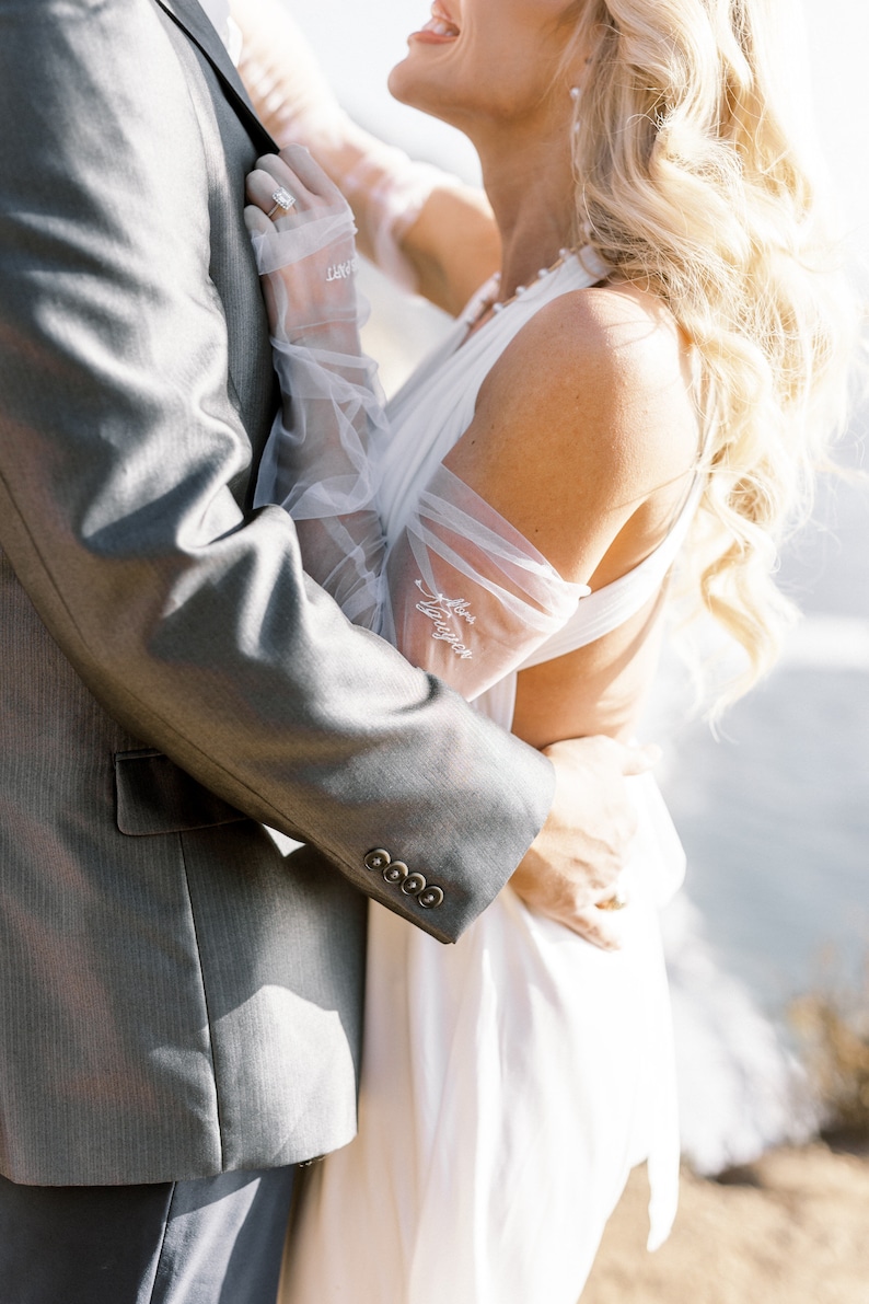 May include: A wedding scene featuring a gray suit jacket with four buttons and a white dress. The bride wears sheer gloves with the text "Mrs. Longton" embroidered on them. A diamond ring is visible. The background is blurred.