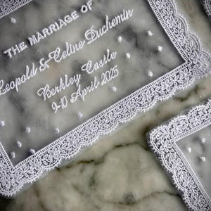 May include: White lace wedding invitation with the text "THE MARRIAGE OF Leopold & Celine Duchemin Berkley Castle 9-10 April 2025". The invitation has a delicate floral lace border and small white polka dots.