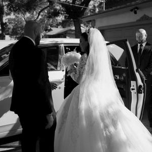 K&ouml;nnte beinhalten: Eine Braut in einem wei&szlig;en Hochzeitskleid mit langem Schleier steigt aus einem wei&szlig;en Auto aus. Ein Mann im schwarzen Anzug hilft ihr. Ein weiterer Mann im schwarzen Anzug steht an der offenen Autot&uuml;r. Die Braut h&auml;lt einen Strau&szlig; wei&szlig;er Blumen.