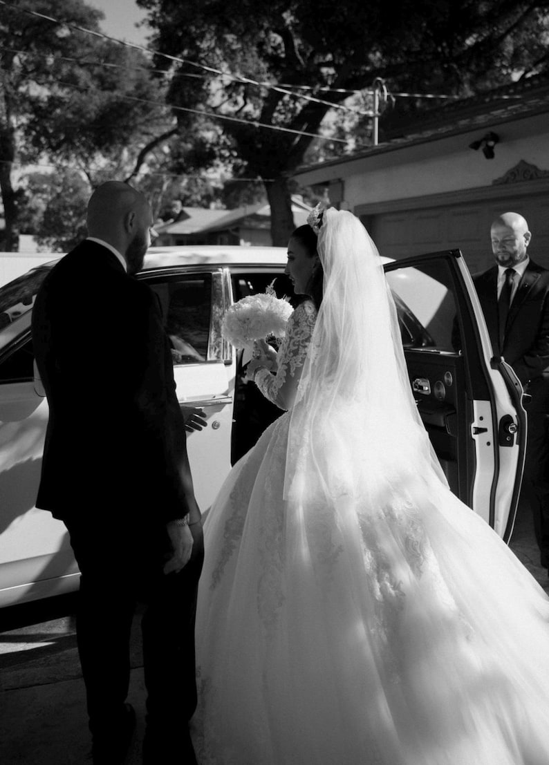 K&ouml;nnte beinhalten: Eine Braut in einem wei&szlig;en Hochzeitskleid mit langem Schleier steigt aus einem wei&szlig;en Auto aus. Ein Mann im schwarzen Anzug hilft ihr. Ein weiterer Mann im schwarzen Anzug steht an der offenen Autot&uuml;r. Die Braut h&auml;lt einen Strau&szlig; wei&szlig;er Blumen.