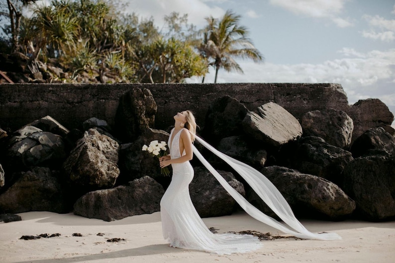 beach destination wedding with bride walking with head tilted back and two long cape shoulder wings blowing in the wind