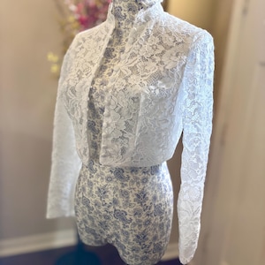 May include: A white lace bolero jacket with long sleeves and a high collar is displayed on a mannequin. The jacket is buttoned in the front. The mannequin is covered in a blue and white floral pattern.