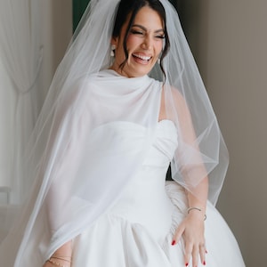 May include: A bride in a white wedding dress and veil laughs. The strapless gown has a full skirt. The veil is draped over the head and shoulders. The bride wears earrings and bracelets. Her nails are painted red.