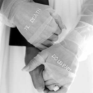 May include: Black and white image of two hands clasped together. The hands are wearing sheer, white gloves with the phrases "TIL DEATH" and "DO US PART" embroidered in white. A diamond ring is visible on one finger.