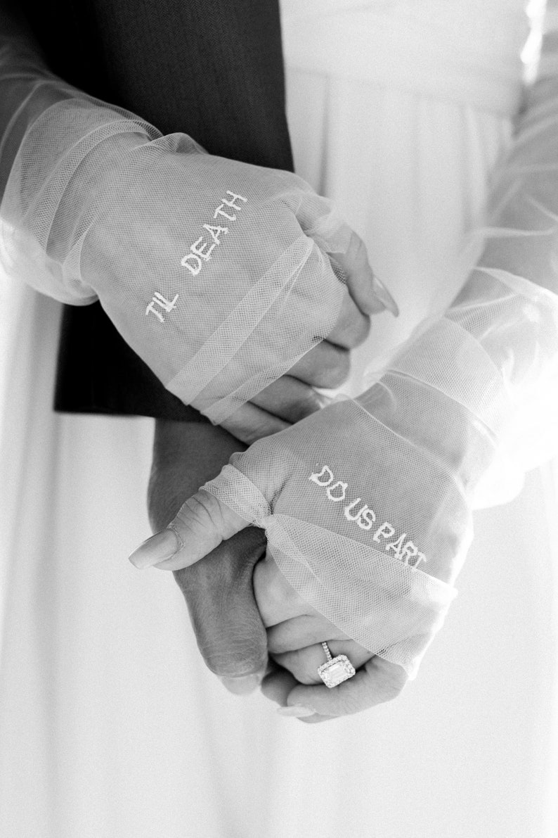 May include: Black and white image of two hands clasped together. The hands are wearing sheer, white gloves with the phrases "TIL DEATH" and "DO US PART" embroidered in white. A diamond ring is visible on one finger.