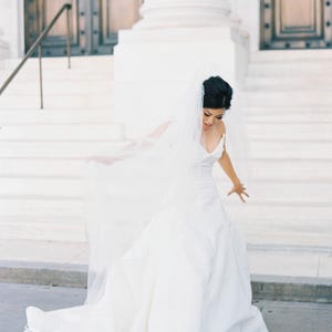 K&ouml;nnte beinhalten: Eine Braut in einem wei&szlig;en Hochzeitskleid mit einem flie&szlig;enden Schleier. Das Kleid hat ein schlichtes Design. Die Braut steht auf wei&szlig;en Stufen vor einem Geb&auml;ude mit gro&szlig;en Holzt&uuml;ren.