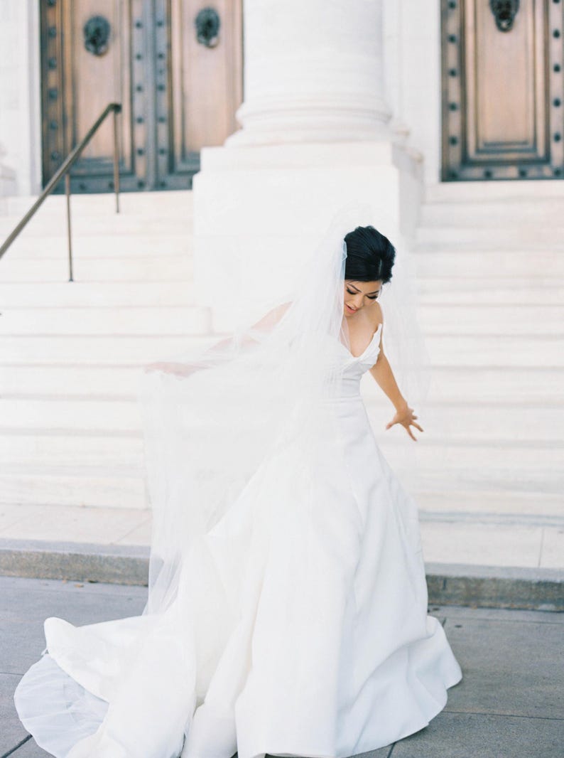 K&ouml;nnte beinhalten: Eine Braut in einem wei&szlig;en Hochzeitskleid mit einem flie&szlig;enden Schleier. Das Kleid hat ein schlichtes Design. Die Braut steht auf wei&szlig;en Stufen vor einem Geb&auml;ude mit gro&szlig;en Holzt&uuml;ren.