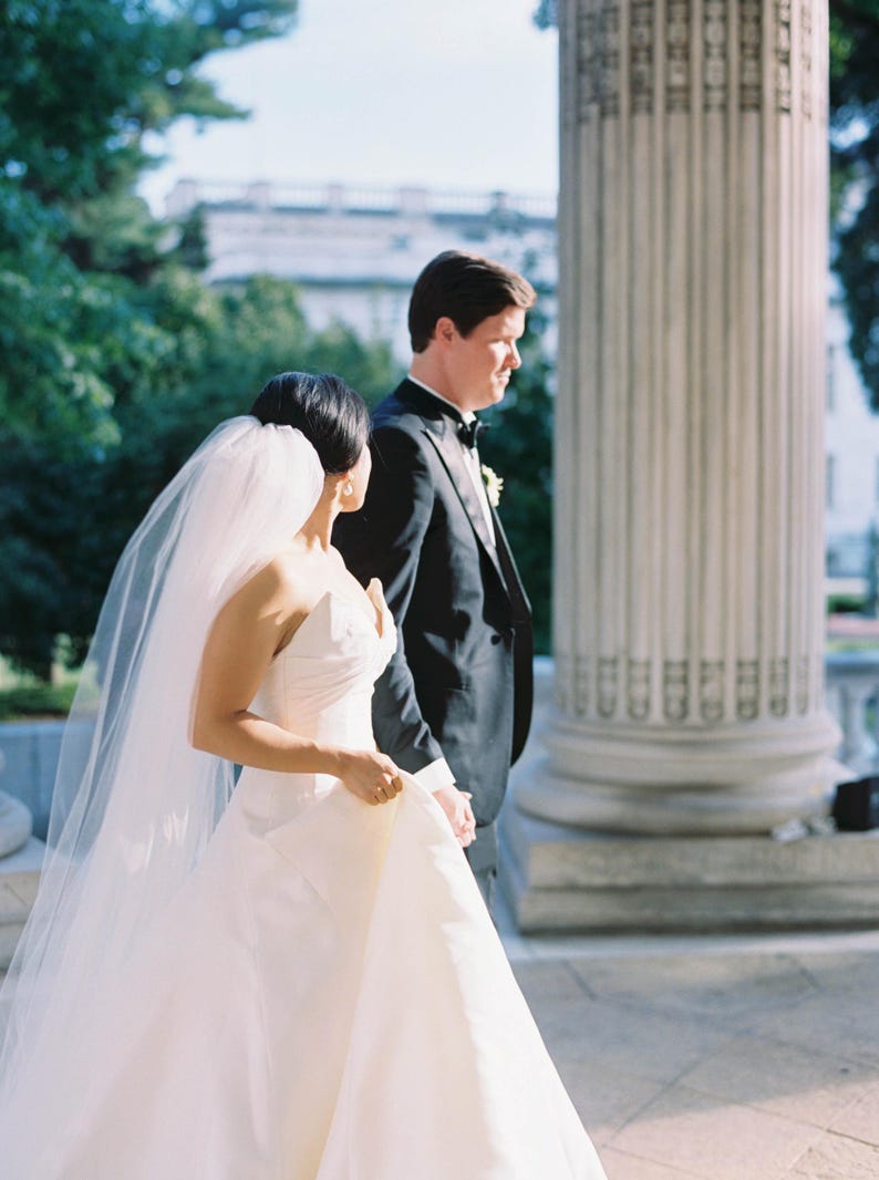 K&ouml;nnte beinhalten: Ein Paar in Hochzeitskleidung geht im Freien. Die Person links tr&auml;gt ein wei&szlig;es Brautkleid und einen Schleier. Die Person rechts tr&auml;gt einen schwarzen Smoking und eine Fliege. Sie halten H&auml;ndchen.