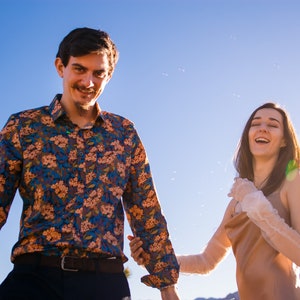 May include: A man wearing a floral print button-down shirt and a woman in a dress and lace gloves are smiling. Bubbles float in the air against a clear blue sky. The couple is holding hands.