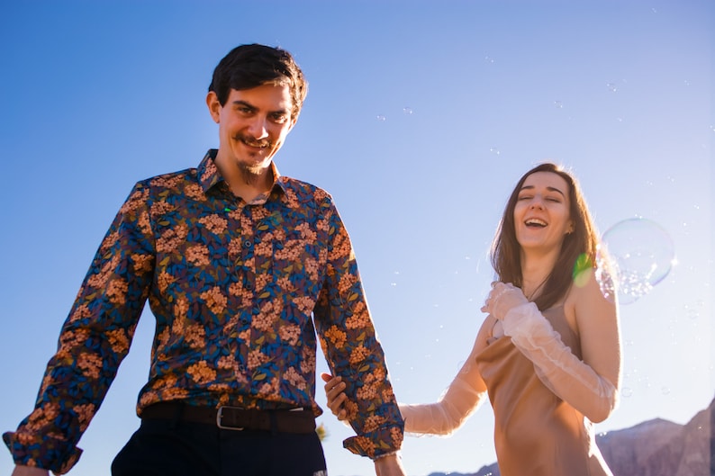 May include: A man wearing a floral print button-down shirt and a woman in a dress and lace gloves are smiling. Bubbles float in the air against a clear blue sky. The couple is holding hands.