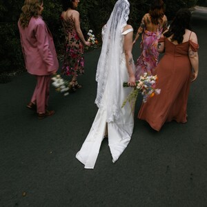 K&ouml;nnte beinhalten: Eine Gruppe von Menschen geht einen Weg entlang, die Person in der Mitte tr&auml;gt ein wei&szlig;es Hochzeitskleid mit Schleier, die anderen Personen tragen bunte Kleider und einen rosa Anzug.