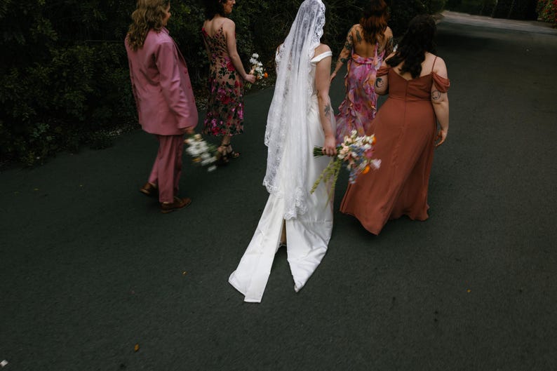 K&ouml;nnte beinhalten: Eine Gruppe von Menschen geht einen Weg entlang, die Person in der Mitte tr&auml;gt ein wei&szlig;es Hochzeitskleid mit Schleier, die anderen Personen tragen bunte Kleider und einen rosa Anzug.