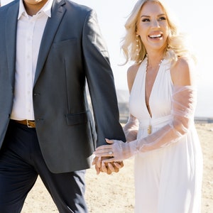 May include: A couple walking outdoors, holding hands. The woman wears a white halter dress and sheer gloves. The man wears a gray suit with a white shirt. The scene is bathed in sunlight, suggesting a wedding or special occasion.