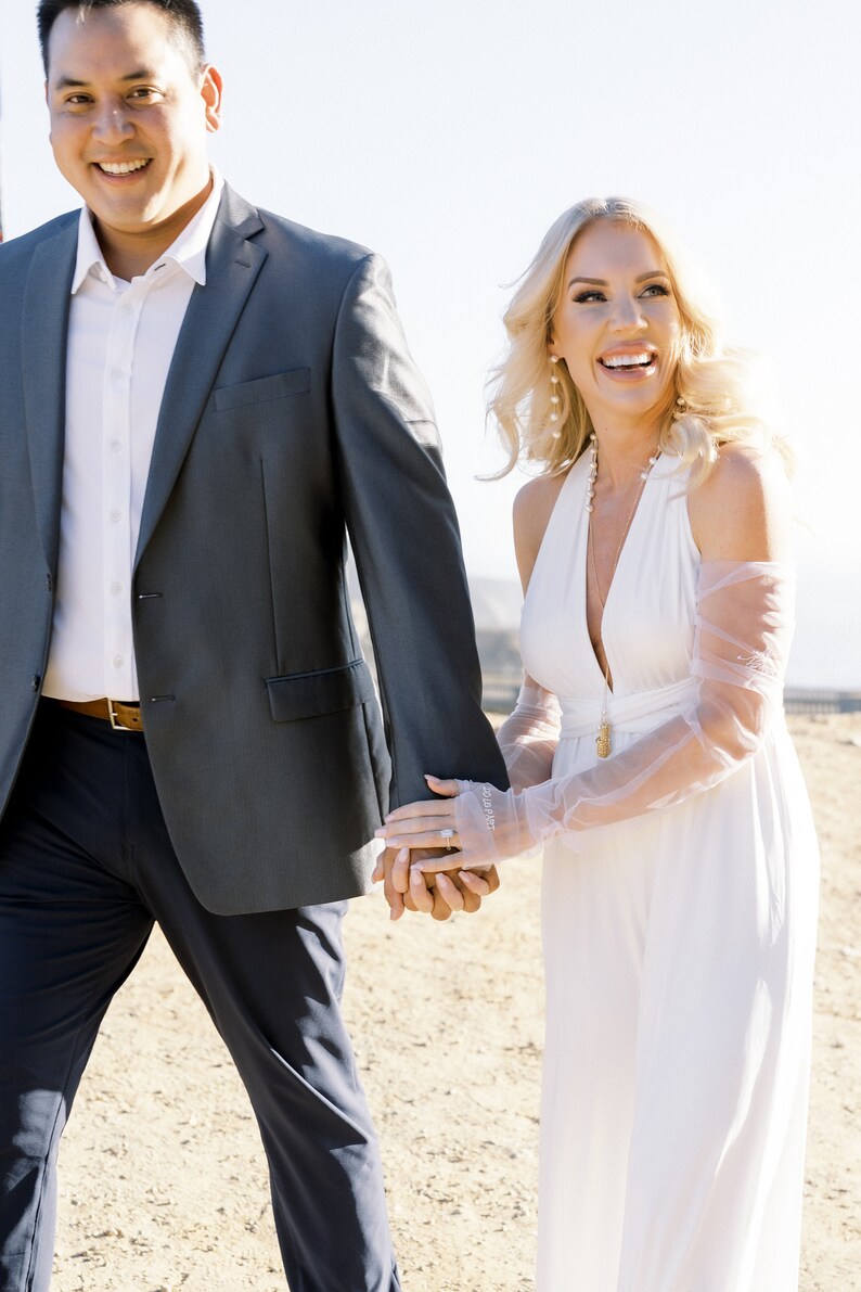 May include: A couple walking outdoors, holding hands. The woman wears a white halter dress and sheer gloves. The man wears a gray suit with a white shirt. The scene is bathed in sunlight, suggesting a wedding or special occasion.