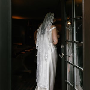 K&ouml;nnte beinhalten: Eine Braut in einem wei&szlig;en Satin-Hochzeitskleid mit einem Spitzen-Schleier. Sie steht in einem T&uuml;rrahmen und schaut auf etwas au&szlig;erhalb des Bildes.