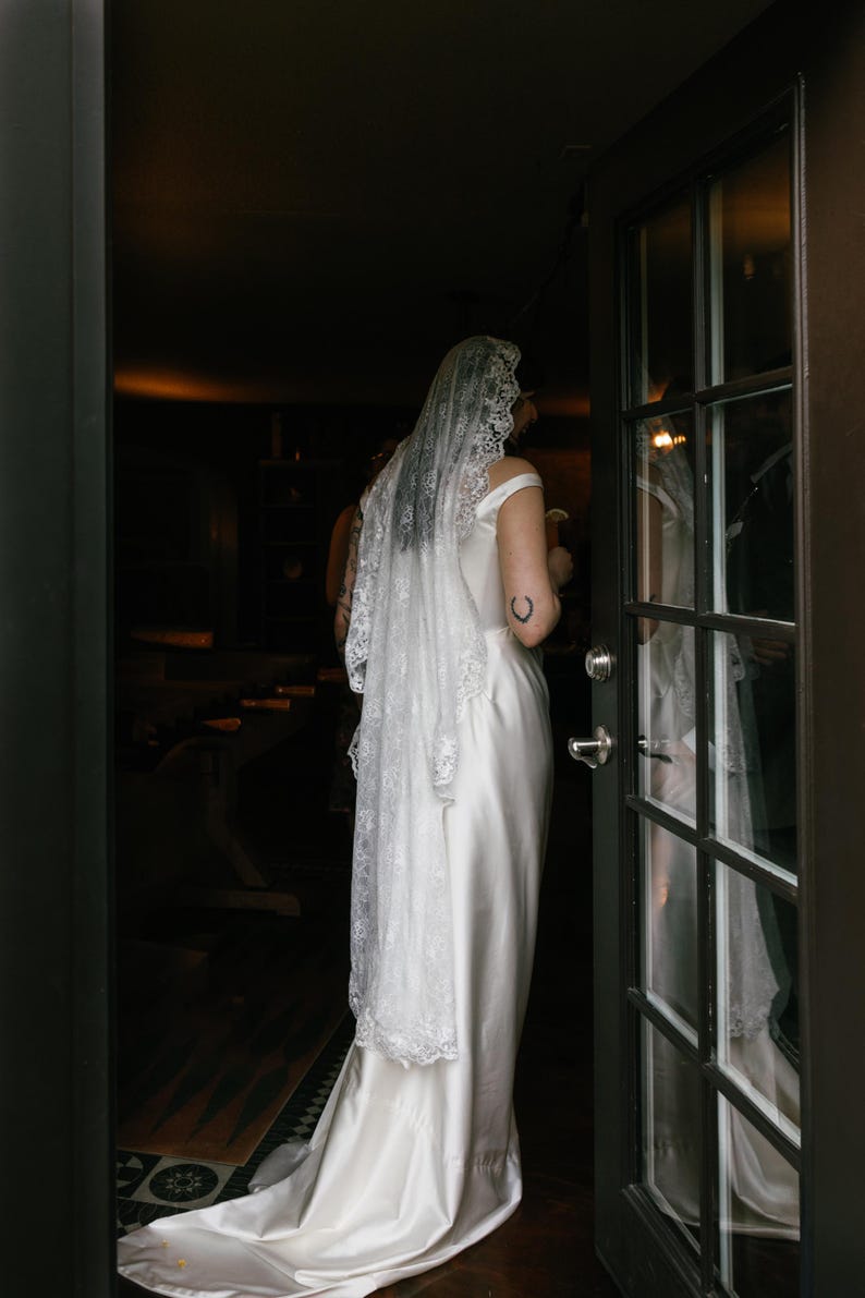 K&ouml;nnte beinhalten: Eine Braut in einem wei&szlig;en Satin-Hochzeitskleid mit einem Spitzen-Schleier. Sie steht in einem T&uuml;rrahmen und schaut auf etwas au&szlig;erhalb des Bildes.