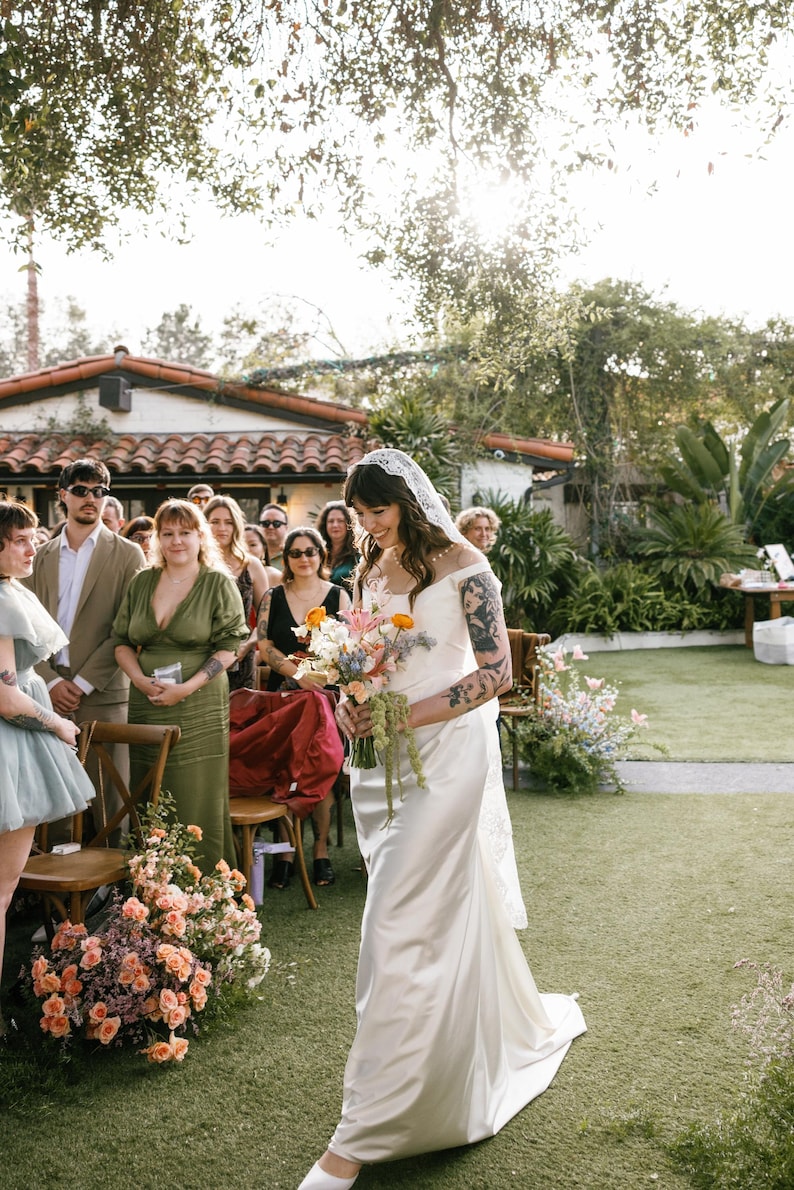 May include: A bride in a white satin dress and a white lace veil walks down the aisle. She is carrying a bouquet of flowers with orange, yellow, and blue flowers. The bride is walking on a green lawn with a group of people seated in chairs behind her.