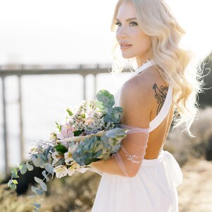 May include: A woman in a white halter dress holds a bouquet of flowers. The bouquet includes white, pink, and green foliage. The woman has blonde hair and a tattoo on her upper back. The words "Mrs. Duggan" are written on her arm.