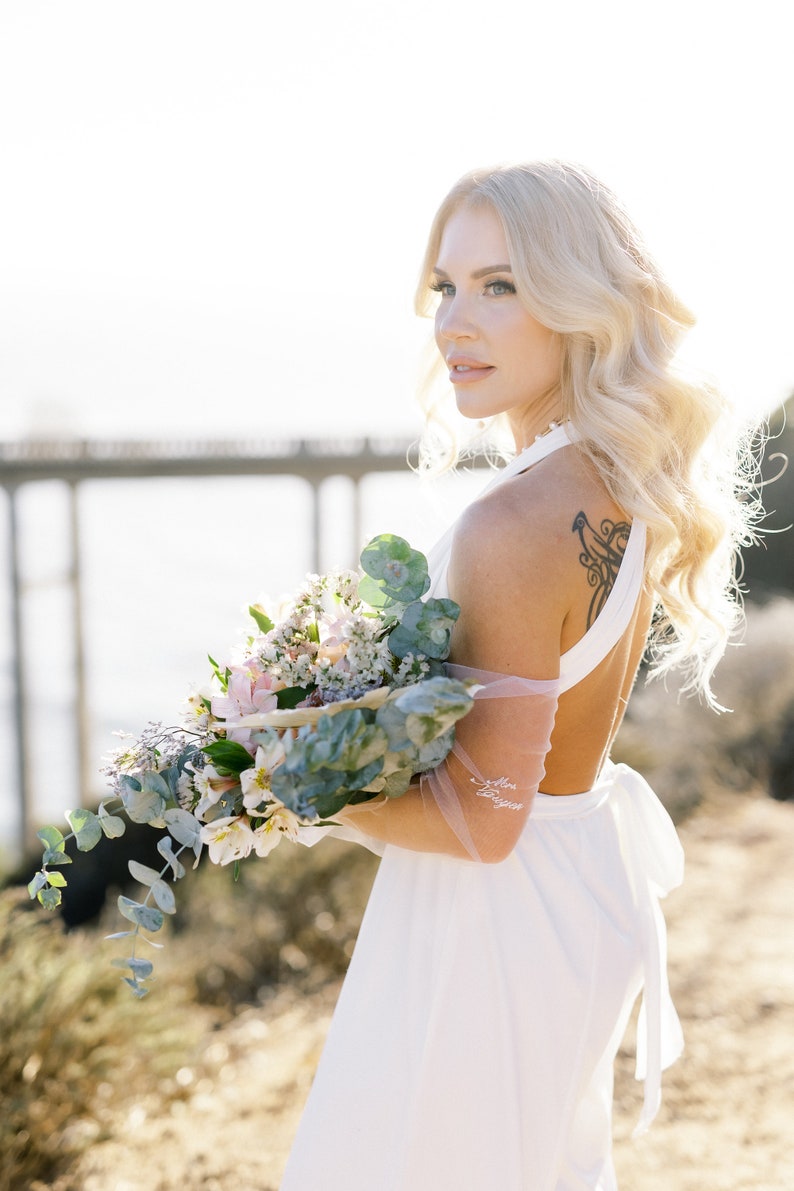 May include: A woman in a white halter dress holds a bouquet of flowers. The bouquet includes white, pink, and green foliage. The woman has blonde hair and a tattoo on her upper back. The words "Mrs. Duggan" are written on her arm.