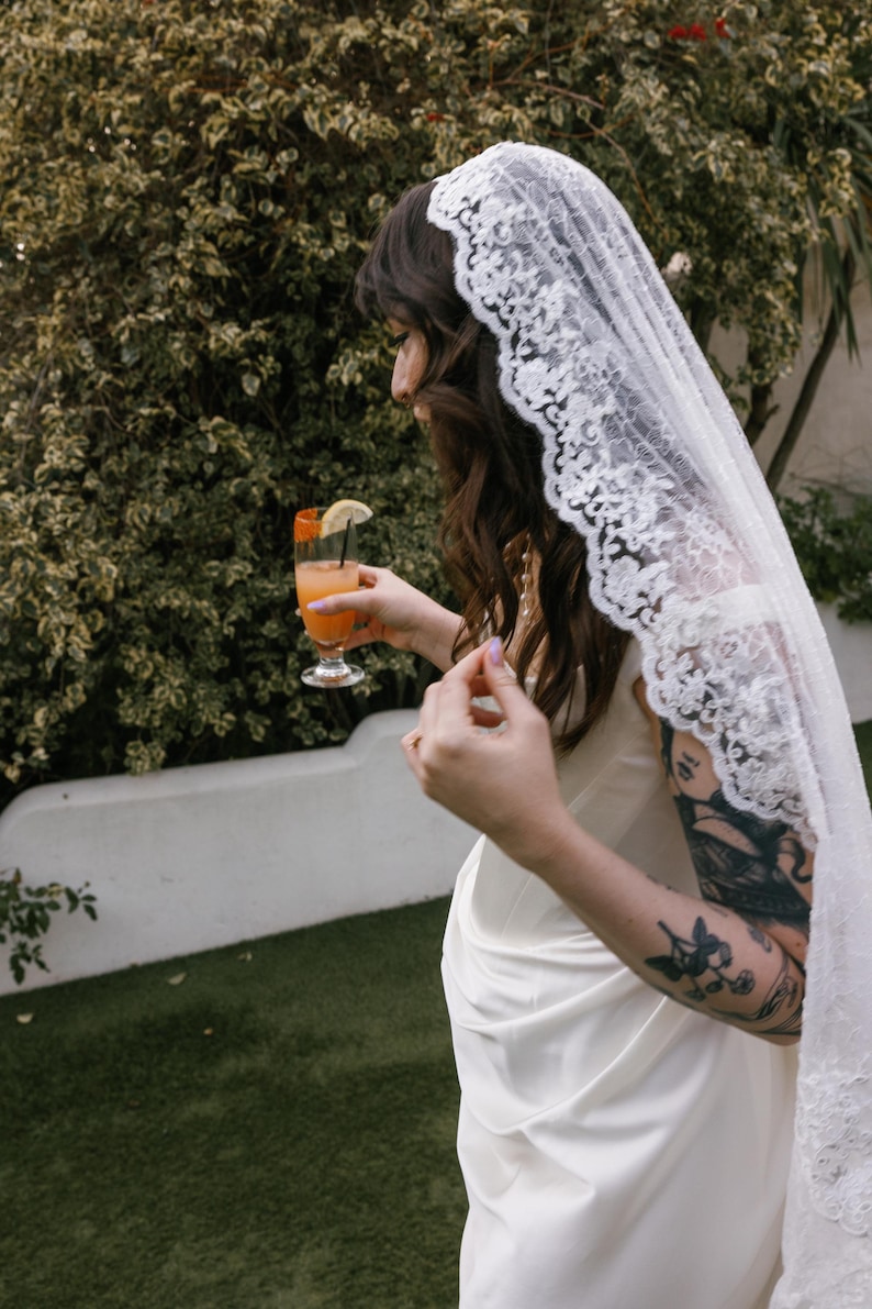 K&ouml;nnte beinhalten: Eine Frau in einem wei&szlig;en Spitzenkleid und einem wei&szlig;en Schleier h&auml;lt ein Glas Orangensaft mit einer Zitronenscheibe. Sie hat ein Tattoo auf ihrem Arm.