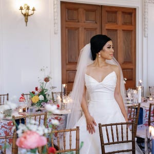 K&ouml;nnte beinhalten: Eine Braut in einem wei&szlig;en, tr&auml;gerlosen Hochzeitskleid und Schleier steht in der N&auml;he eines Tisches, der f&uuml;r einen Hochzeitsempfang gedeckt ist. Der Tisch ist mit Blumenarrangements und Kerzen dekoriert. Holzst&uuml;hle sind um den Tisch angeordnet.