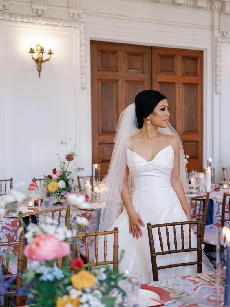 K&ouml;nnte beinhalten: Eine Braut in einem wei&szlig;en, tr&auml;gerlosen Hochzeitskleid und Schleier steht in der N&auml;he eines Tisches, der f&uuml;r einen Hochzeitsempfang gedeckt ist. Der Tisch ist mit Blumenarrangements und Kerzen dekoriert. Holzst&uuml;hle sind um den Tisch angeordnet.