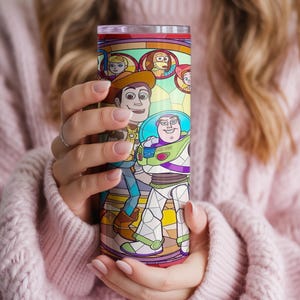 Puede incluir: Un vaso alto y colorido con un dise&ntilde;o de estilo vidriera con personajes de la pel&iacute;cula animada "Toy Story". El dise&ntilde;o incluye a Woody, Buzz Lightyear y otros personajes. El vaso es sostenido por una persona que lleva un su&eacute;ter rosa.