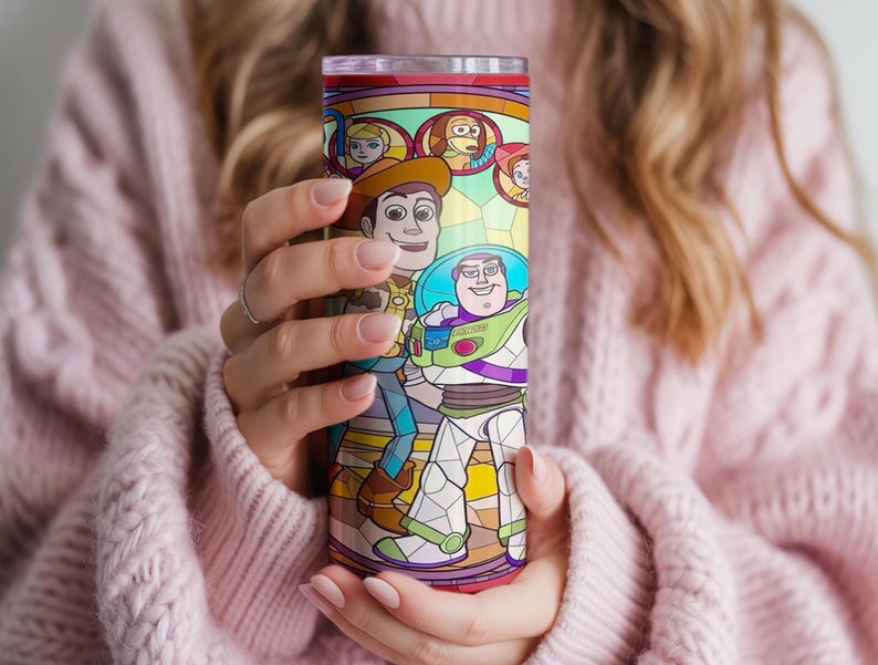 Puede incluir: Un vaso alto y colorido con un dise&ntilde;o de estilo vidriera con personajes de la pel&iacute;cula animada "Toy Story". El dise&ntilde;o incluye a Woody, Buzz Lightyear y otros personajes. El vaso es sostenido por una persona que lleva un su&eacute;ter rosa.